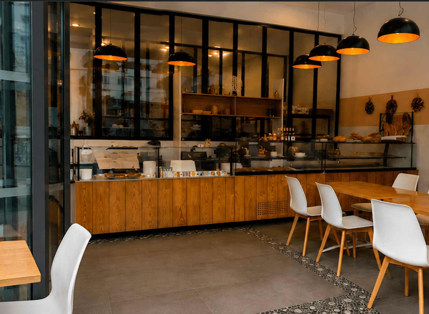 Intérieur d'une boulangerie moderne avec comptoirs en bois, chaises blanches et suspensions noires élégantes.
