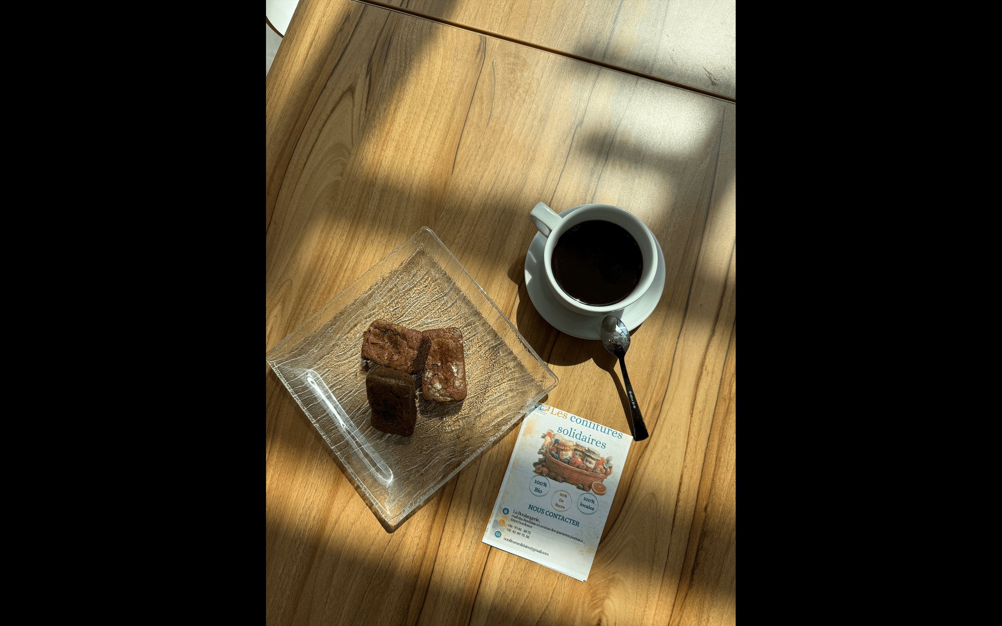 Café noir, brownies sur assiette en verre et dépliant posés sur une table en bois.