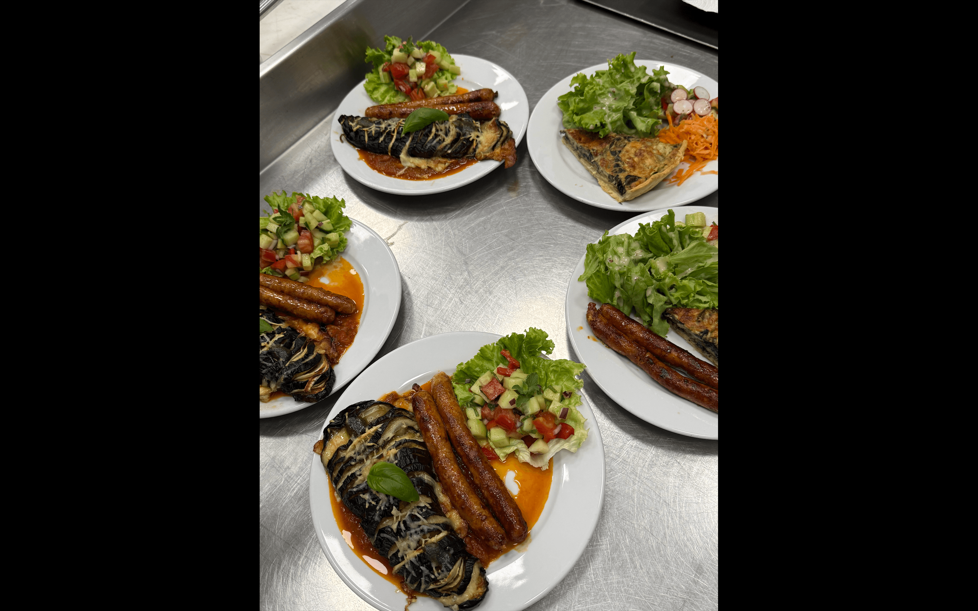 Cinq assiettes avec saucisses, aubergines gratinées, quiche et salades fraîches sur un comptoir métallique.