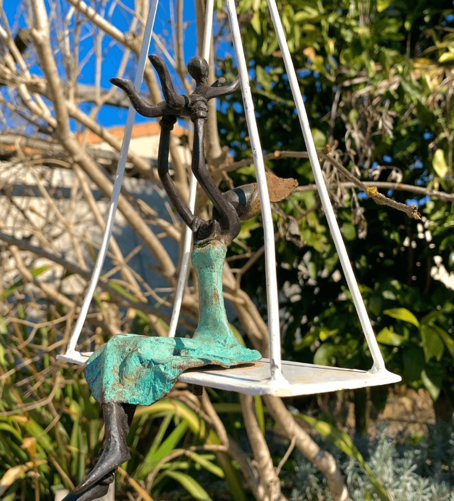 Petite sculpture d'une femme aux bras levés assise sur une balançoire blanche en plein air.