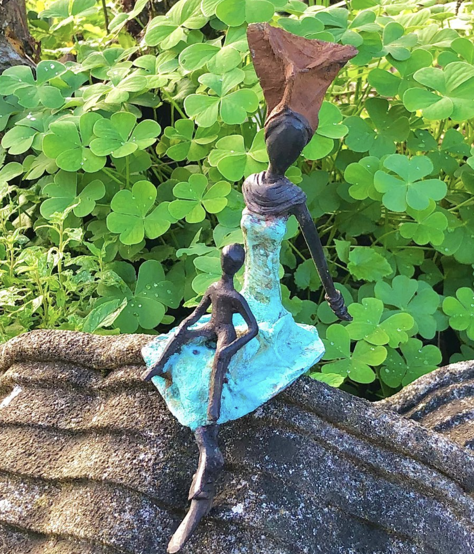Petite sculpture d'une femme en robe turquoise et d'un enfant devant des trèfles verts.